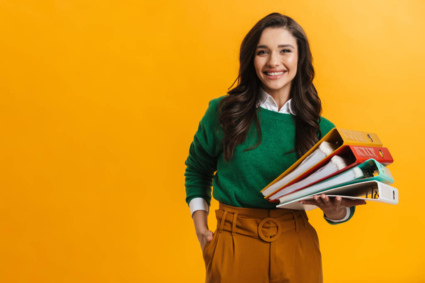 Portrait of a happy smiling casual woman standing over yellow background holding stack of office folders - Foto, Imagen