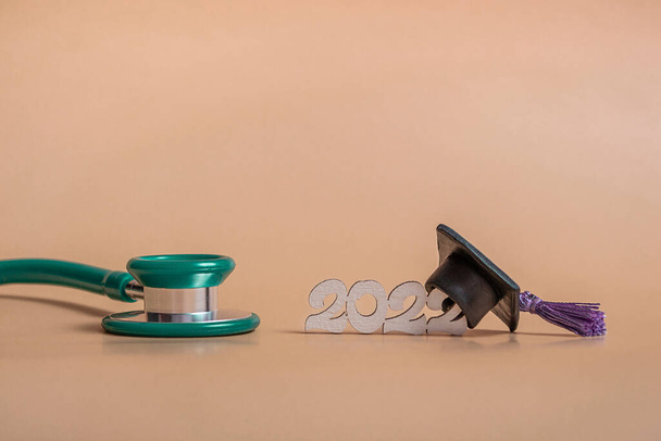 Absolventenmütze mit Stethoskop und Ziffern 2022 auf beigem Hintergrund, Nahaufnahme mit Kopierraum. Medizinisches Ausbildungskonzept. - Foto, Bild