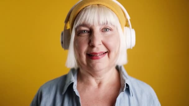 Carefree blonde hair 50s woman with closed eyes dancing to the rhythm of the melody, listening to the song in wireless headphones in studio - Felvétel, videó