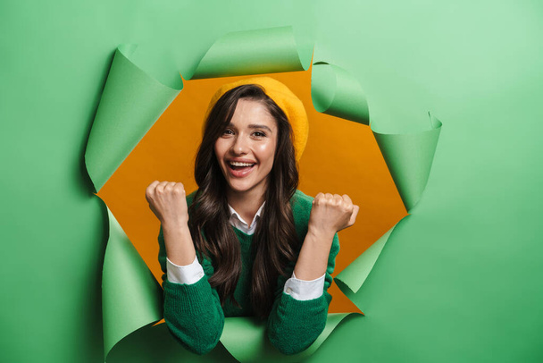 Young white smiling woman peeking out hole isolated on green background celebrating success - Фото, изображение