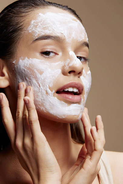 retrato mujer crema en la mejilla posando cosméticos para limpiar la piel de fondo beige - Foto, Imagen