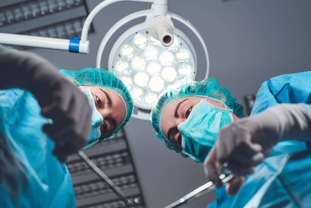 Alhaalta naispuoliset kirurgit lääketieteellisessä univormussa käyttäen ammattimaisia työkaluja seisoessaan kirkkaassa valossa leikkaussalissa - Valokuva, kuva