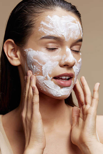 όμορφη γυναίκα φροντίδα του δέρματος με τη χρήση λευκής μάσκας στο πρόσωπο μπεζ φόντο - Φωτογραφία, εικόνα