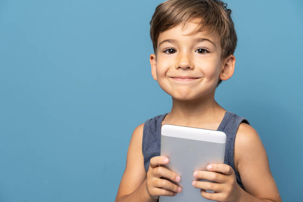 Dört yaşında, mavi arka plan stüdyosunun önünde dikilip dijital tablet kullanarak kameraya gülümseyen küçük beyaz bir çocuğun ön görüntüsü. - Fotoğraf, Görsel