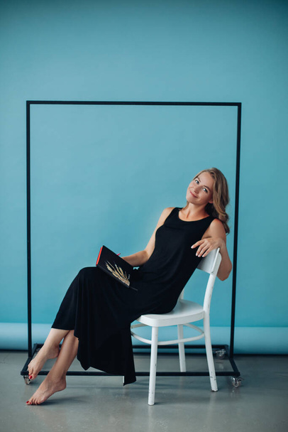 One attractive smiling Caucasian model in long black dress sitting on white chair leaning on its back holding planner in hand and smiling at camera against clothing rack on blue background. - Фото, изображение