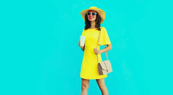 Retrato de hermosa mujer feliz sonriente con taza de café con sombrero de verano amarillo y vestido sobre fondo azul - Foto, imagen