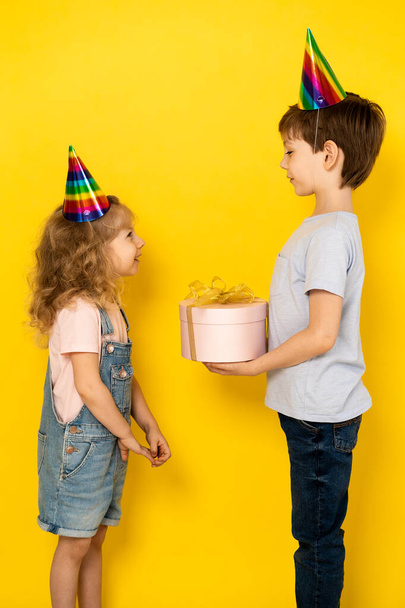 Küçük sarışın kız, erkek kardeşinden doğum günü hediyesi olarak pembe kutuda hediye kabul eder. Sarı arka planda, şapkalı çocuklar, dikey fotoğraf. - Fotoğraf, Görsel