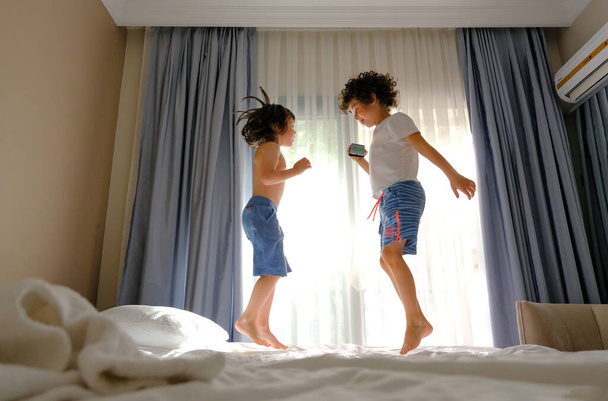 Dwoje szczęśliwych dzieciaków skaczących na łóżko z telefonem słuchającym muzyki - Zdjęcie, obraz