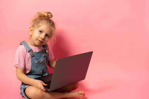 Uśmiechnięta dziewczynka siedzi z komputerowym laptopem odizolowanym na różowym tle. Kształcenie na odległość w domu podczas kwarantanny. baner z miejscem na tekst - Zdjęcie, obraz