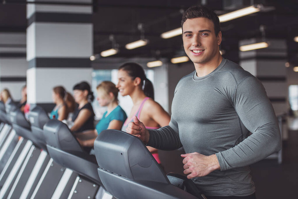 Attractive young muscular man running on a treadmill in gym, looking at camera and smiling - Fotó, kép