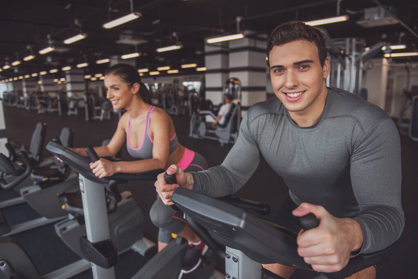 Привлекательные молодые мускулистые люди тренируются на велотренажере в спортзале и улыбаются
 - Фото, изображение