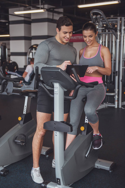 Attractive young woman working out on an exercise bike in gym, muscular trainer consulting her. Both smiling - Photo, Image