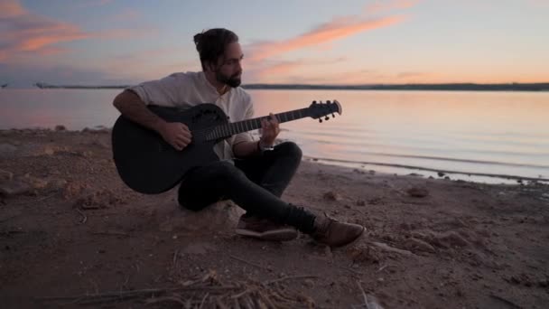 Homme caucasien espagnol jouant de la guitare sur la plage au bord du lac au coucher du soleil  - Séquence, vidéo