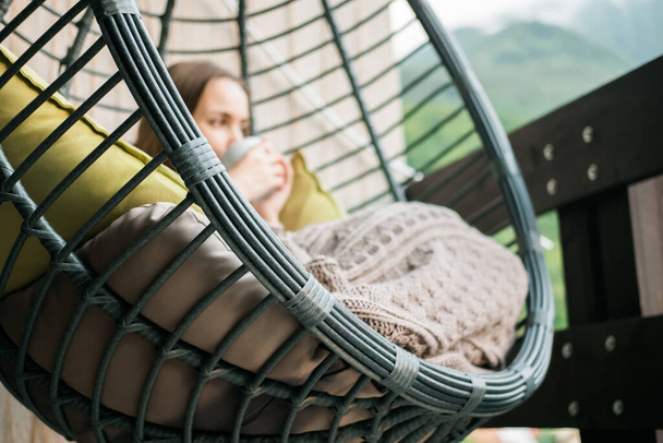 Young woman sitting in a hanging chair with a blanket and a cup of coffee on the balcony. - Foto, imagen
