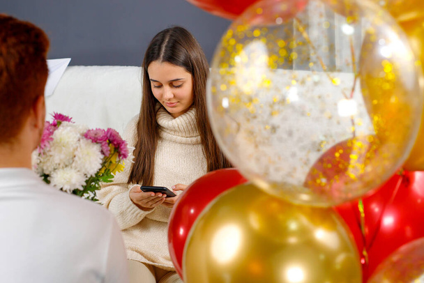 Young man making surprise for his girlfriend holding flowers in his hands and colored balloons while she sitting on a sofa looking at the phone. - Fotografie, Obrázek