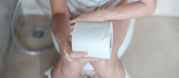 Kobieta trzymająca rolkę papieru toaletowego podczas siedzenia na muszli klozetowej. biegunka, okres zaparcia, zakażenie, brzuch, ból brzucha, zapalenie pęcherza moczowego, koncepcja ubezpieczenia i medycyny - Zdjęcie, obraz