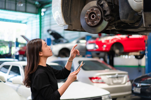 Asian young woman in black shirt examining a vehicle suspension and break system in garage close up with copyspace. Woman inspecting a car on a hydraulic ramp in garage. - Foto, Imagem