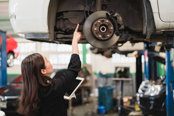Asian young woman in black shirt examining a vehicle suspension and break system in garage close up with copyspace. Woman inspecting a car on a hydraulic ramp in garage. - Foto, immagini