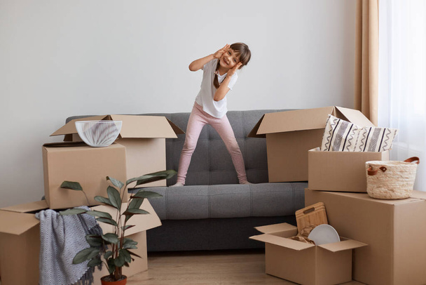 Portrait of happy excited little girl wearing casual style attire standing on sofa and having fun, posing surrounded with boxes of belongings, relocating, moving to a new apartment. - Foto, imagen
