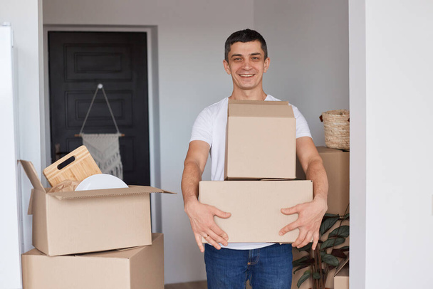 Portrait of happy satisfied man wearing white T-shirt standing with cardboard boxes with belongings, unpacking packages with stuff, looking at camera with smile and positive expression. - Photo, Image