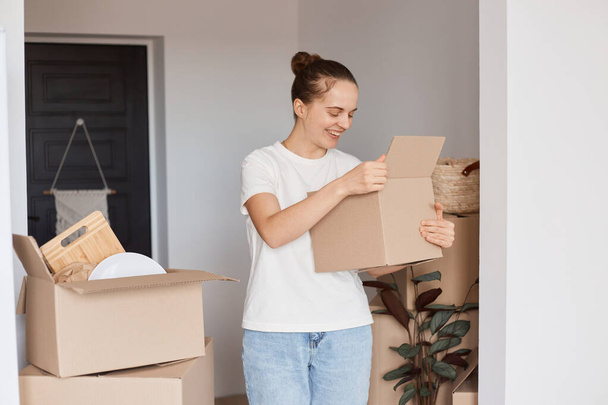 Portrait of beautiful Caucasian woman wearing white casual style T-shirt and jeans standing holding cardboard box with stuff, looking inside a package, unpacking belongings. - Foto, Bild