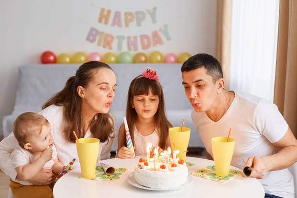 Happy cute female child making wish and going to blow candles on cake while celebrating Birthday with family, sitting at table with birthday decoration on background. - Фото, изображение