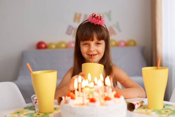 Indoor shot of dark haired smiling little sitting at table with cake and candles, drinking lemonade from yellow glasses, expressing happy positive emotions, looking at camera. - Фото, изображение
