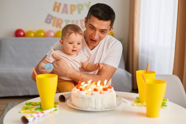 Indoor shot of father and toddler child with birthday cake sitting at table and looking at delicious dessert, celebrating birthday, blowing candles, posing at home with sofa and balloons on background - Photo, image