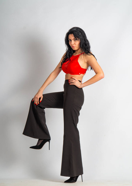 atractiva modelo morena joven sexy usando en top rojo y pantalones negros posando en la cámara en la habitación blanca - Foto, imagen