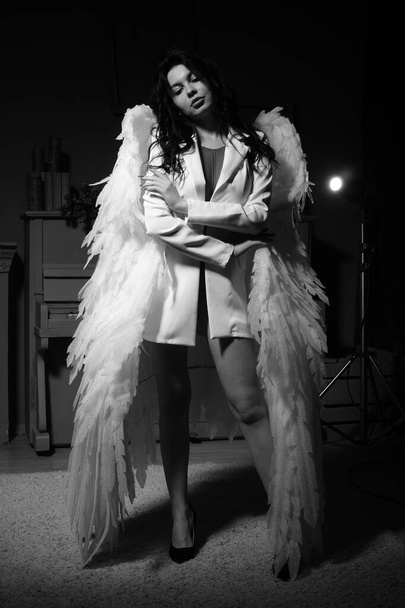 Mooi sexy meisje in lingerie en jas met vleugels poseren bij de camera in de studio, Zwart-wit foto - Foto, afbeelding
