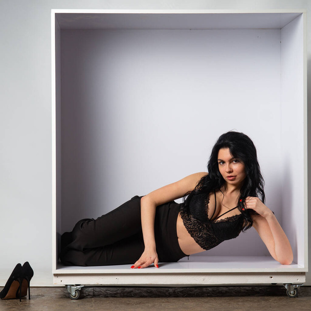 ελκυστική σέξι νεαρή μελαχρινή μοντέλο φοράει σε μαύρο crop top και μαύρο παντελόνι ποζάρουν στην κάμερα στο λευκό δωμάτιο - Φωτογραφία, εικόνα