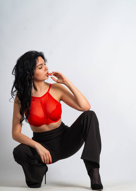 привлекательная сексуальная молодая брюнетка в красном топе и черные брюки позируют перед камерой в белой комнате - Фото, изображение