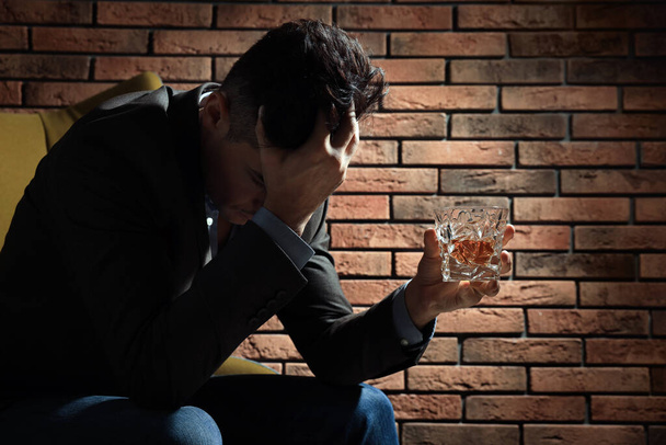 Addicted man with glass of alcoholic drink in armchair near red brick wall - 写真・画像