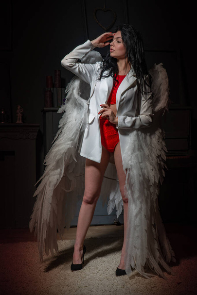 Piękna seksowna dziewczyna w czerwonej bieliźnie i białej kurtce ze skrzydłami pozującymi do kamery w studio  - Zdjęcie, obraz