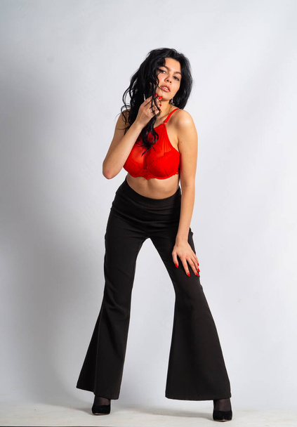 vonzó szexi fiatal barna modell visel piros termés felső és fekete nadrág pózol a kamera a fehér szobában - Fotó, kép
