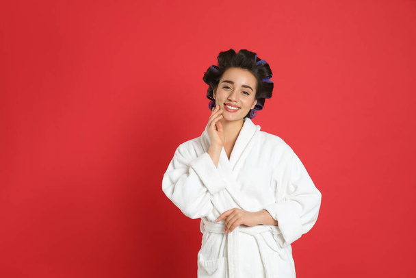 Happy young woman in bathrobe with hair curlers on red background, space for text - Foto, afbeelding