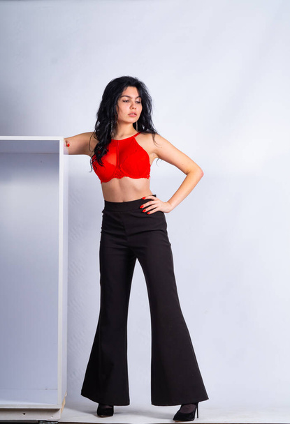 atractiva modelo morena joven sexy usando en top rojo y pantalones negros posando en la cámara en la habitación blanca - Foto, Imagen
