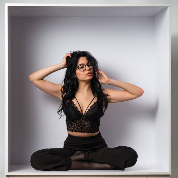 atraente sexy jovem morena modelo vestindo em preto cultura top e preto calças posando para a câmera no branco quarto - Foto, Imagem