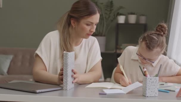 Střední pomalý záběr maminky a dcery dělat dívky domácí úkoly spolu sedí za stolem v útulném obývacím pokoji - Záběry, video