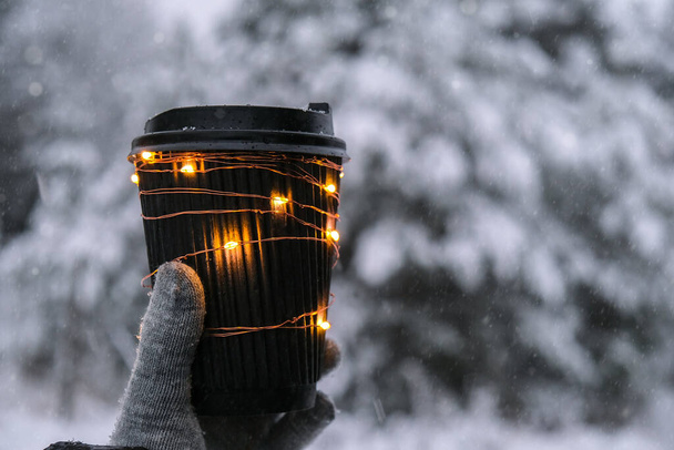 Žena ruka v zimě teplé rukavice drží černý eko papírový šálek s girland lesklé světlo. Tvůrčí módní kelímek s nulovým odpadem. Mockup. Horký ohřívací nápoj jít v zimním dni. Odnést koncepci volného času koncept čaj nebo šálek kávy - Fotografie, Obrázek
