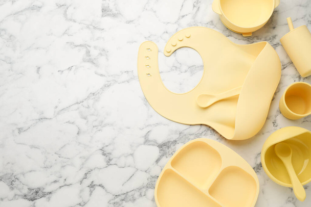 Flat lay composition with silicone baby bib and plastic dishware on white marble background. Space for text - Фото, зображення