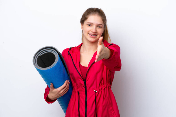 Teenager girl going to yoga classes while holding a mat isolated on white bakcground shaking hands for closing a good deal - Photo, Image