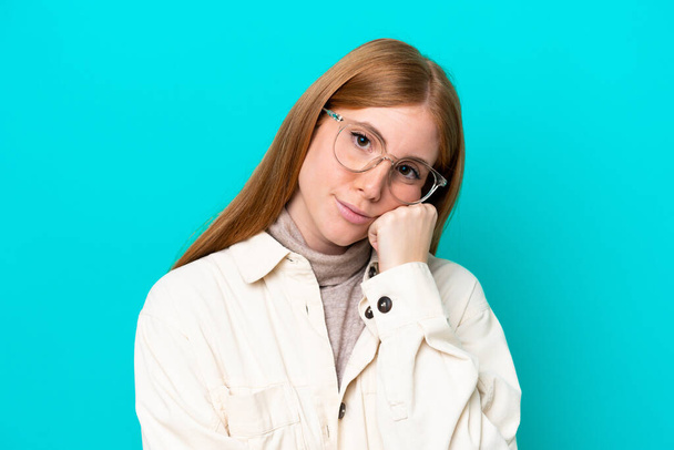 Giovane donna rossa isolata su sfondo blu con occhiali e con espressione triste - Foto, immagini