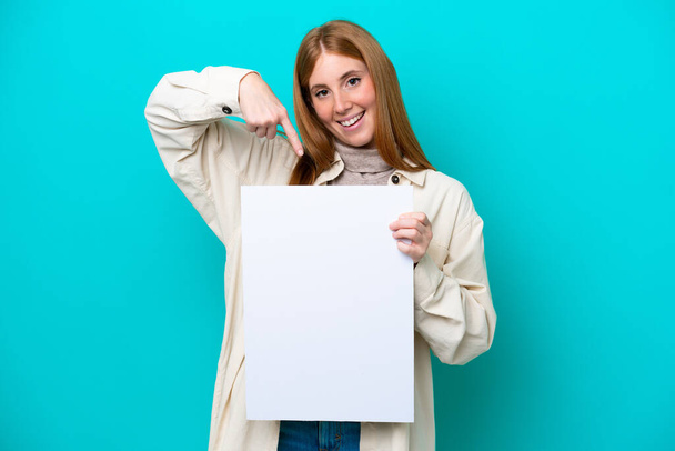 Fiatal vörös hajú nő elszigetelt kék háttér kezében egy üres plakát boldog kifejezést, és rámutatva rá - Fotó, kép