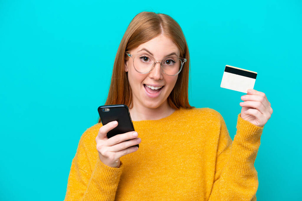 Młoda ruda kobieta odizolowana na niebieskim tle kupuje za pomocą telefonu komórkowego i trzyma kartę kredytową o zaskakującym wyrazie twarzy - Zdjęcie, obraz