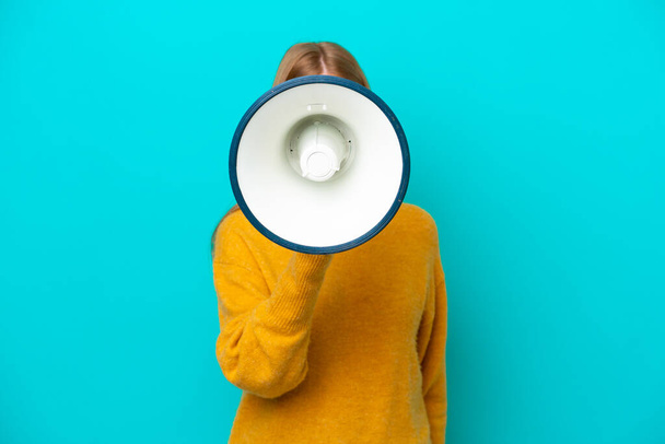 Young redhead woman isolated on blue background shouting through a megaphone to announce something - Photo, image