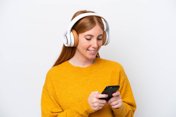 Genç kızıl saçlı kadın beyaz arka planda izole edilmiş müzik dinliyor ve cep telefonu arıyor. - Fotoğraf, Görsel