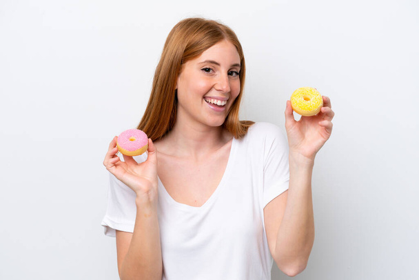Genç kızıl saçlı kadın beyaz arka planda izole edilmiş elinde mutlu bir ifadeyle donut tutuyor. - Fotoğraf, Görsel