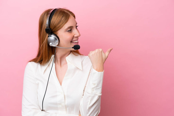 Telemarketer redhead woman working with a headset isolated on pink background pointing to the side to present a product - Photo, image