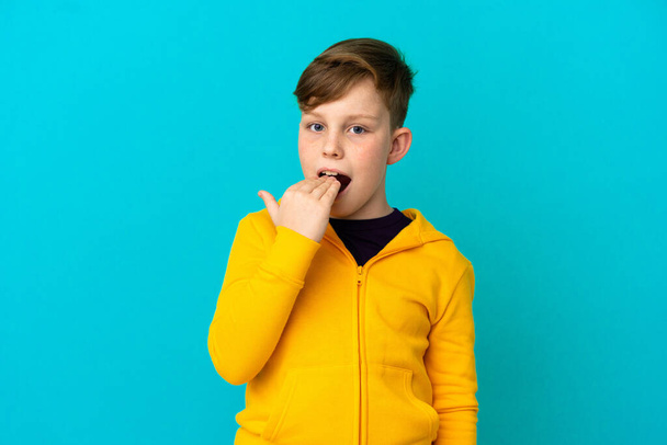 Μικρό κοκκινομάλλικο αγόρι απομονωμένο σε μπλε φόντο χασμουριέται και καλύπτει το ανοιχτό στόμα με το χέρι - Φωτογραφία, εικόνα
